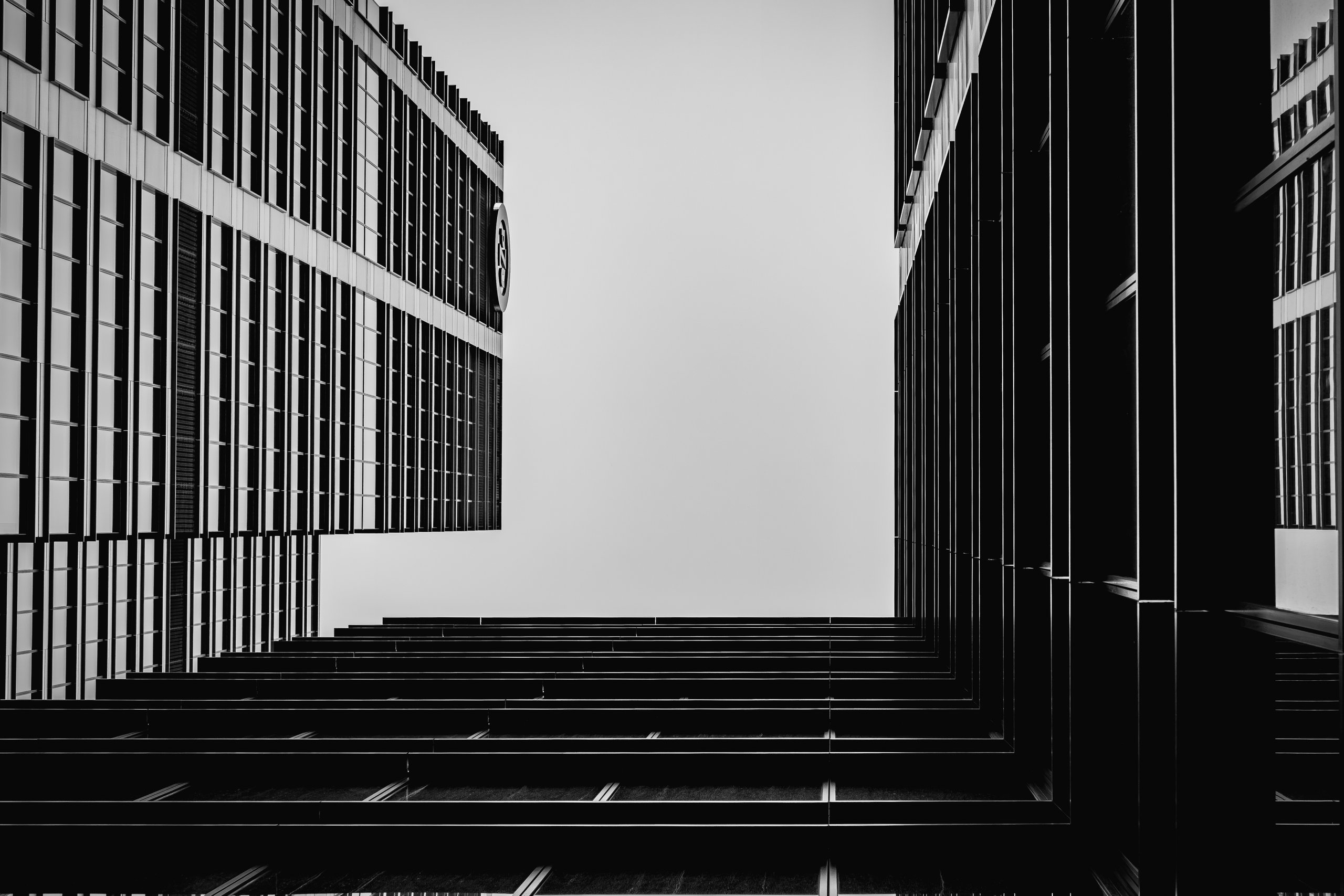 Two office facades converging on a pale sky, looking straight up between them in black and white